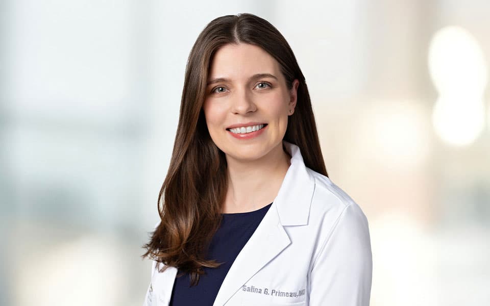 Dr Primeau wearing a white lab coat, smiling against a softly blurred light background.