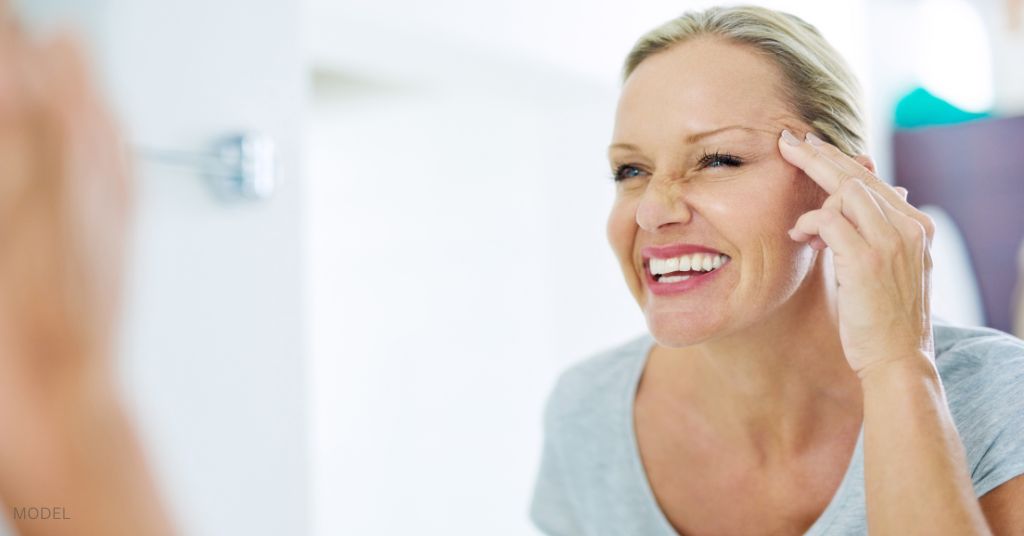 Woman looking in the mirror and smiling while touching her face. (MODEL)