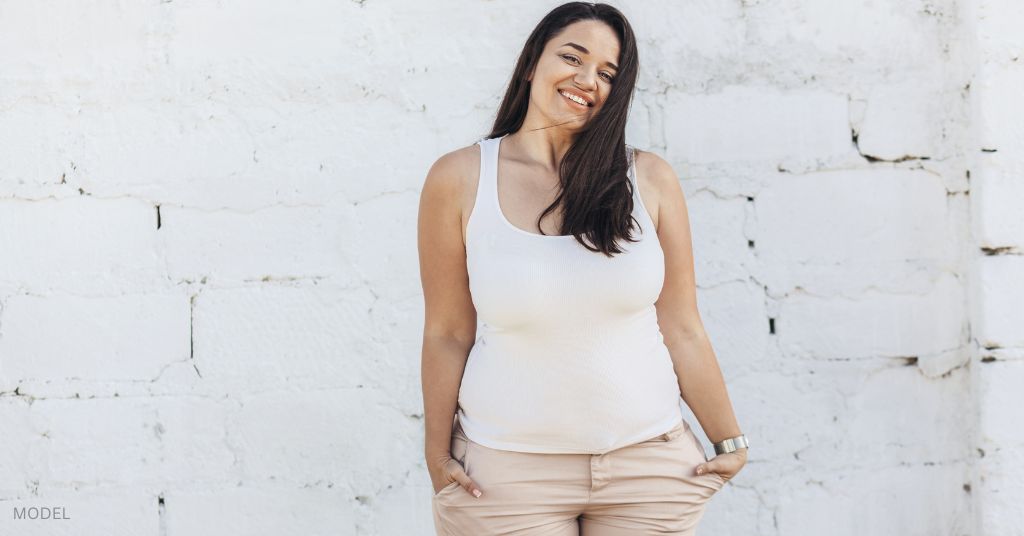 Woman standing with her hands in her pockets smiling. (MODEL)