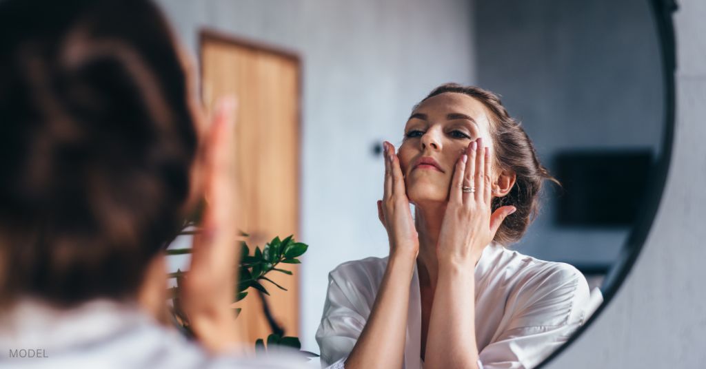 A woman looks at her reflection in the mirror while holding her face.